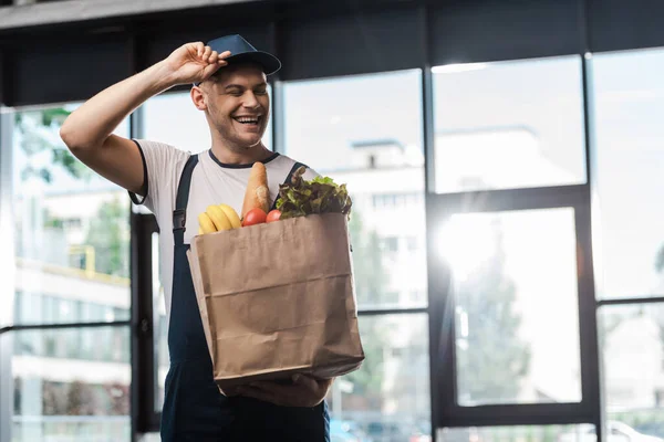Happy delivery man touching cap and holding paper bag with groceries — Stock Photo