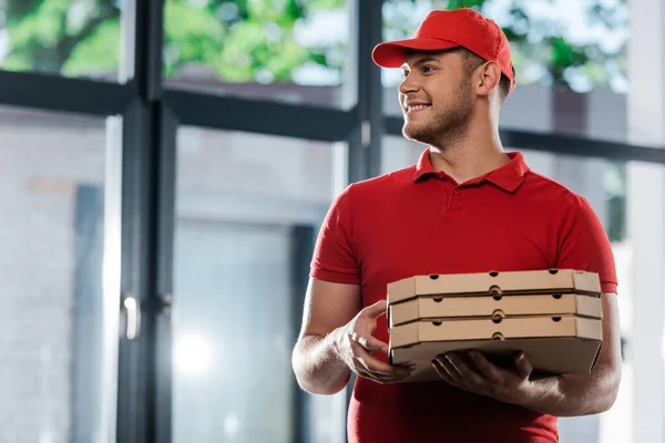 Glücklicher Zusteller mit Mütze schaut weg und hält Pizzakartons in der Hand — Stockfoto