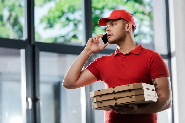 Guapo repartidor en gorra hablando en el teléfono inteligente y sosteniendo cajas de pizza - foto de stock