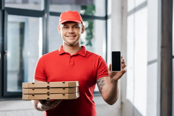Felice uomo di consegna in cap con smartphone con schermo bianco e scatole di pizza — Foto stock