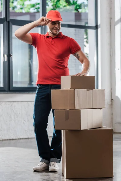 Cheerful delivery man touching cap and looking at carton boxes — Stock Photo