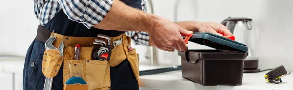 Panoramic crop of plumber in tool belt opening toolbox on worktop in kitchen — Stock Photo