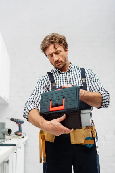 Guapo fontanero en overoles sosteniendo caja de herramientas en la cocina - foto de stock