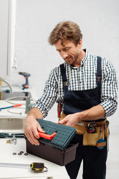 Selective focus of plumber in workwear opening toolbox near tools on worktop in kitchen — Stock Photo