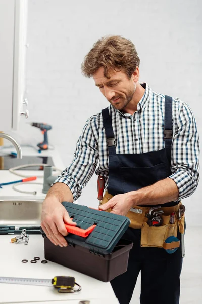 Enfoque selectivo del fontanero en la caja de herramientas de apertura de ropa de trabajo cerca de herramientas en la encimera en la cocina - foto de stock