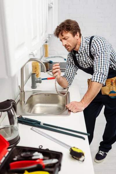 Foco seletivo de encanador em macacão segurando tubo de metal perto de pia na cozinha — Fotografia de Stock