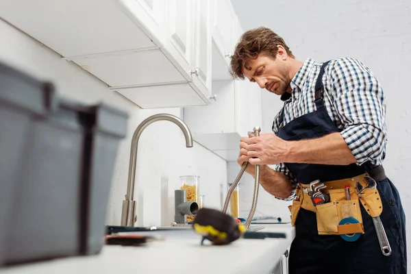 Concentration sélective du plombier tenant le tuyau métallique près du robinet de cuisine et des outils sur le plan de travail — Photo de stock