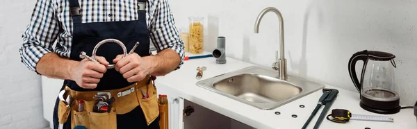 Panoramic crop of plumber in tool belt holding metal pipe in kitchen — Stock Photo