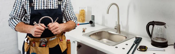 Cultivo panorámico de fontanero en correa de herramientas que sostiene el tubo de metal en la cocina - foto de stock