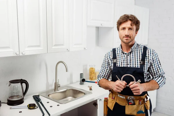 Handsome plumber holding metal pipe and looking at camera in kitchen — Stock Photo