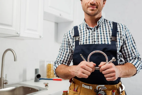 Vista recortada del fontanero en ropa de trabajo y correa de herramientas que sostiene el tubo de metal en la cocina - foto de stock