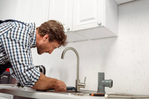 Seitenansicht des Klempners, der einen Rohrschlüssel hält, während er den Küchenhahn in der Nähe von Werkzeugen auf der Arbeitsplatte repariert — Stockfoto