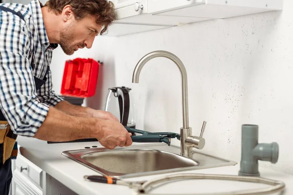 Vue latérale du beau réparateur utilisant une clé à tuyau tout en fixant le robinet de cuisine — Photo de stock