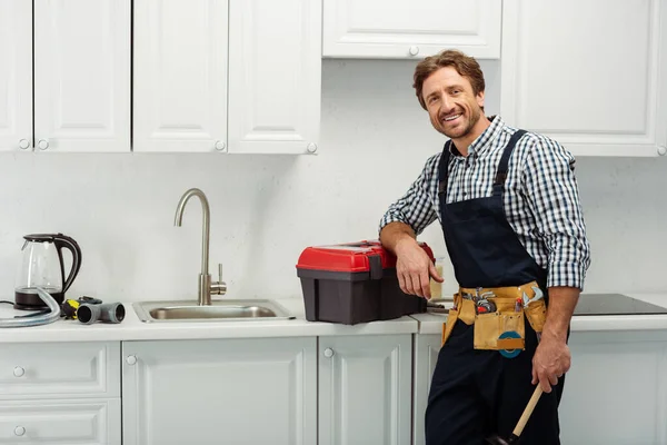 Plombier souriant regardant la caméra tout en tenant le piston près de la boîte à outils sur le plan de travail de la cuisine — Photo de stock