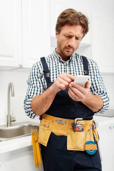 Enfoque selectivo de fontanero guapo en cinturón de herramientas y overoles usando smartphone en cocina - foto de stock