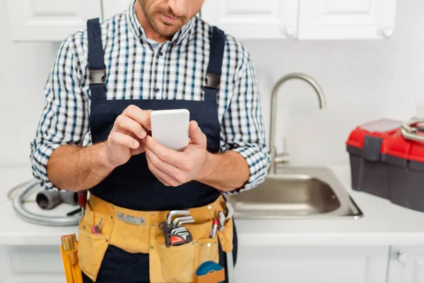 Vista recortada del trabajador en el cinturón de herramientas utilizando el teléfono inteligente mientras trabaja en la cocina - foto de stock