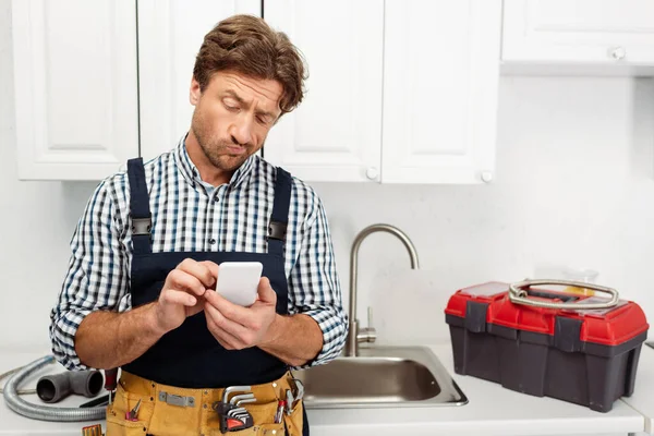 Plomero pensativo con teléfono inteligente cerca de la caja de herramientas y fregadero en la cocina - foto de stock