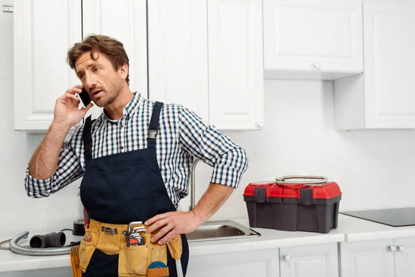 Plombier en vêtements de travail et ceinture d'outils parlant sur smartphone près des outils sur le plan de travail dans la cuisine — Photo de stock