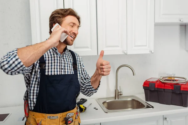 Encanador sorrindo mostrando como gesto e falando no smartphone perto da pia da cozinha — Fotografia de Stock