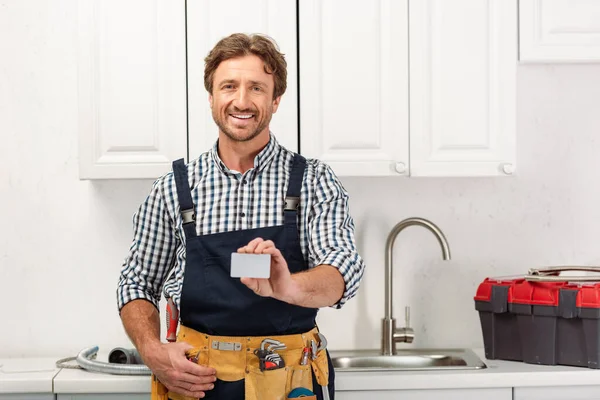 Усміхнений сантехнік в робочому одязі тримає порожню картку і дивиться на камеру на кухні — стокове фото