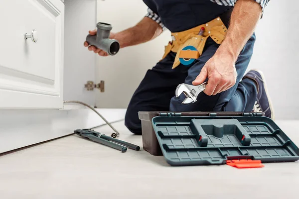 Cropped view of plumber holding plastic pipe and wrench near sink in kitchen — Stock Photo