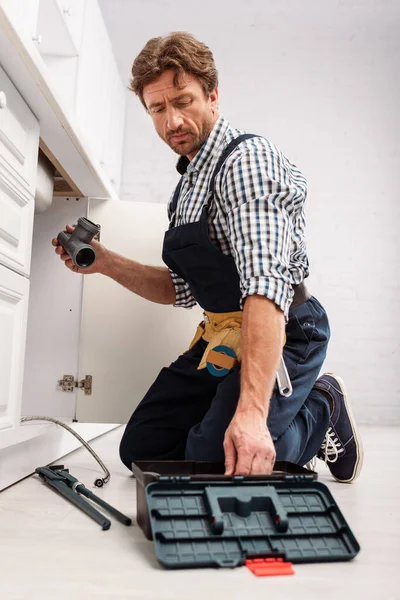 Selektiver Fokus eines gutaussehenden Reparateurs, der Kunststoffrohr in der Nähe von Werkzeugkasten und Rohrschlüssel auf dem Boden in der Küche hält — Stockfoto