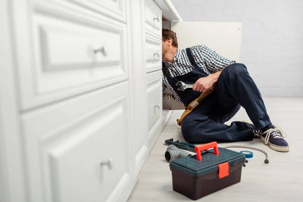 Foco selectivo del fontanero sosteniendo la llave inglesa mientras reparaba el fregadero de la cocina cerca de herramientas y caja de herramientas en el piso - foto de stock