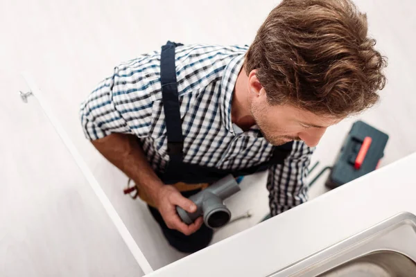 Vista aérea del fontanero sosteniendo tubería de plástico mientras reparaba el fregadero cerca de la caja de herramientas en la cocina - foto de stock