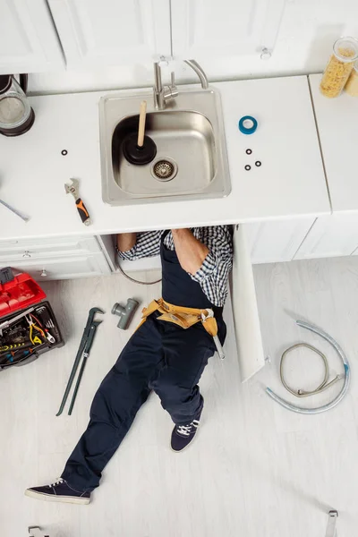 Vista dall'alto dell'idraulico nel lavandino di fissaggio dell'abbigliamento da lavoro vicino agli strumenti sul pavimento — Foto stock