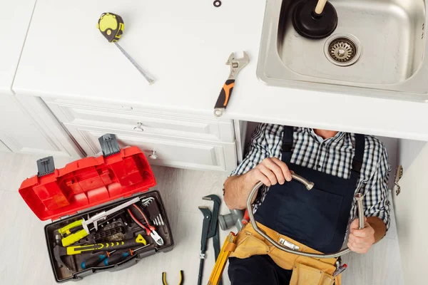 Vue de dessus du plombier tenant le tuyau métallique tout en réparant l'évier de cuisine près des outils sur le plan de travail et le plancher — Photo de stock