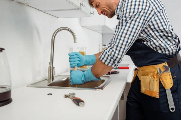 Side view of handsome plumber in tool belt and rubber gloves cleaning kitchen sink with plunger — Stock Photo