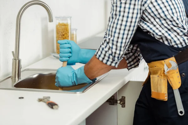 Vue recadrée du plombier dans la ceinture à outils et gants en caoutchouc nettoyage blocage de l'évier de cuisine — Photo de stock