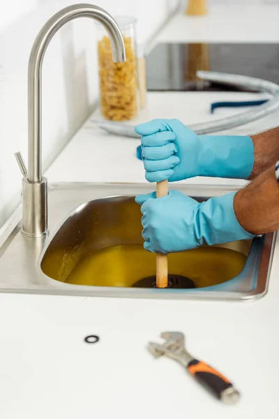 Vista recortada del trabajador en guantes de goma limpieza fregadero de cocina con émbolo cerca de la llave inglesa en la encimera - foto de stock