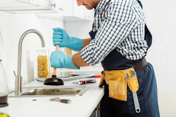 Cropped view of plumber in tool belt and rubber gloves holding plunger near tools on kitchen worktop — Stock Photo