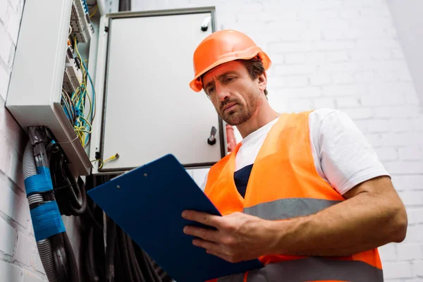 Vista de ángulo bajo del guapo electricista sujetando el portapapeles cerca de la caja de distribución eléctrica - foto de stock