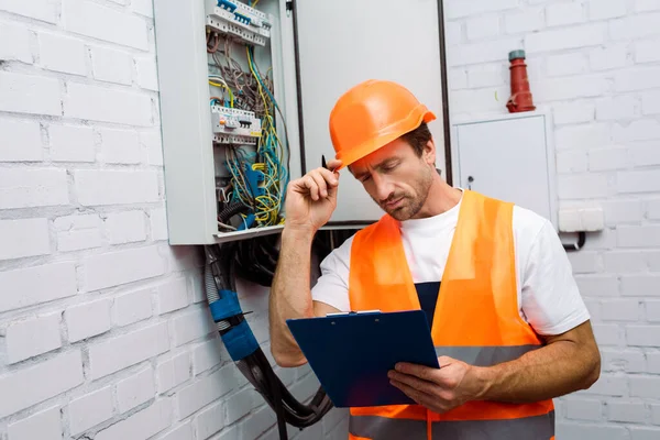 Guapo electricista sujetando portapapeles y bolígrafo mientras trabaja cerca del panel eléctrico - foto de stock