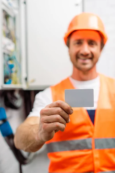 Enfoque selectivo del electricista sonriente en hardhat sosteniendo la tarjeta vacía - foto de stock