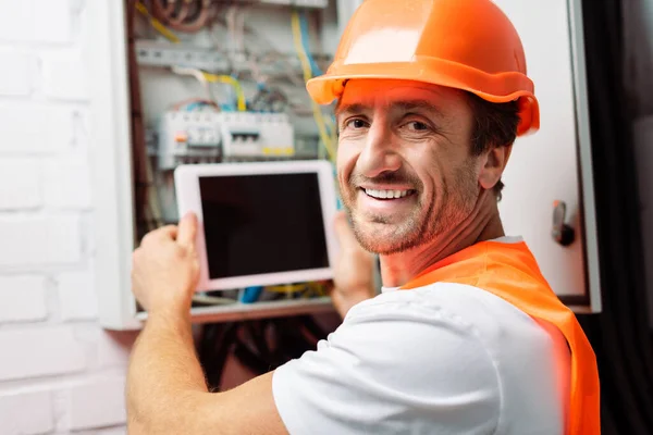 Selective focus of smiling electrician in hardhat holding digital tablet while working with electric panel — Stock Photo