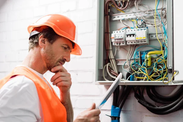 Seitenansicht eines nachdenklichen Elektrikers, der ein digitales Tablet in der Nähe eines Stromverteilers hält — Stockfoto