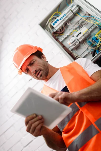 Selektiver Fokus des gut aussehenden Arbeiters in Warnweste und Helm mittels digitalem Tablet in der Nähe von Stromkasten — Stockfoto