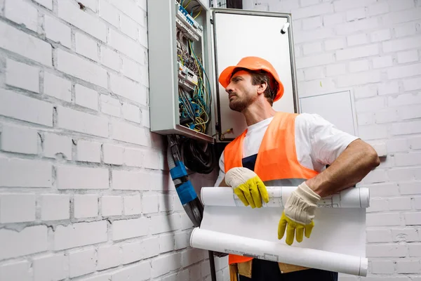 Guapo electricista sosteniendo el plano cerca de la caja de distribución eléctrica - foto de stock