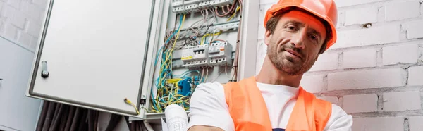 Imagen panorámica del electricista con plano mirando a la cámara cerca de la caja eléctrica - foto de stock