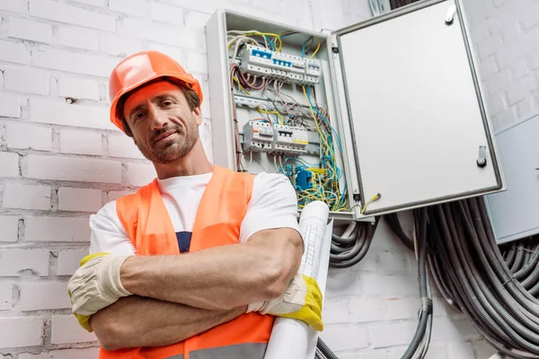 Obrero guapo en casco y chaleco de seguridad sosteniendo el plano y mirando a la cámara cerca del panel eléctrico - foto de stock