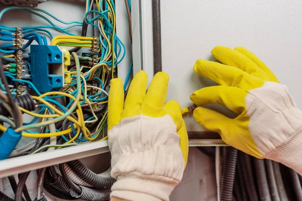 Vista recortada del electricista en guantes sosteniendo alambre cerca del panel eléctrico - foto de stock