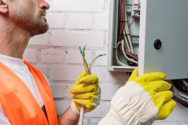 Vista recortada del electricista en guantes sosteniendo cables cerca de la caja de distribución eléctrica - foto de stock