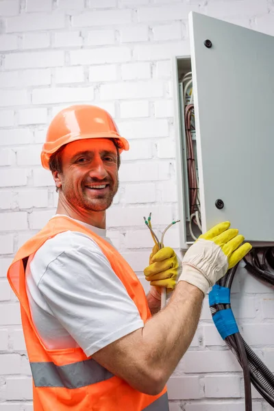 Vista laterale dell'elettricista in hardhat che sorride alla telecamera mentre tiene i fili vicino alla scatola di distribuzione elettrica — Foto stock