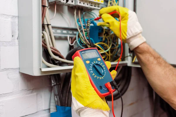 Vista recortada del trabajador con guantes usando multímetro mientras comprueba el voltaje del panel eléctrico - foto de stock
