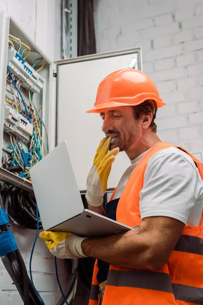 Vista laterale dell'elettricista che toglie il guanto e tiene il computer portatile vicino alla scatola di distribuzione elettrica — Foto stock