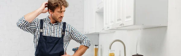 Encanador pensivo com mão perto da cabeça olhando para a torneira na cozinha, orientação panorâmica — Fotografia de Stock