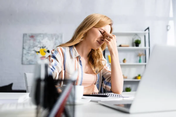 Selective focus of tired freelancer touching eyes near laptop and stationery on table — Stock Photo